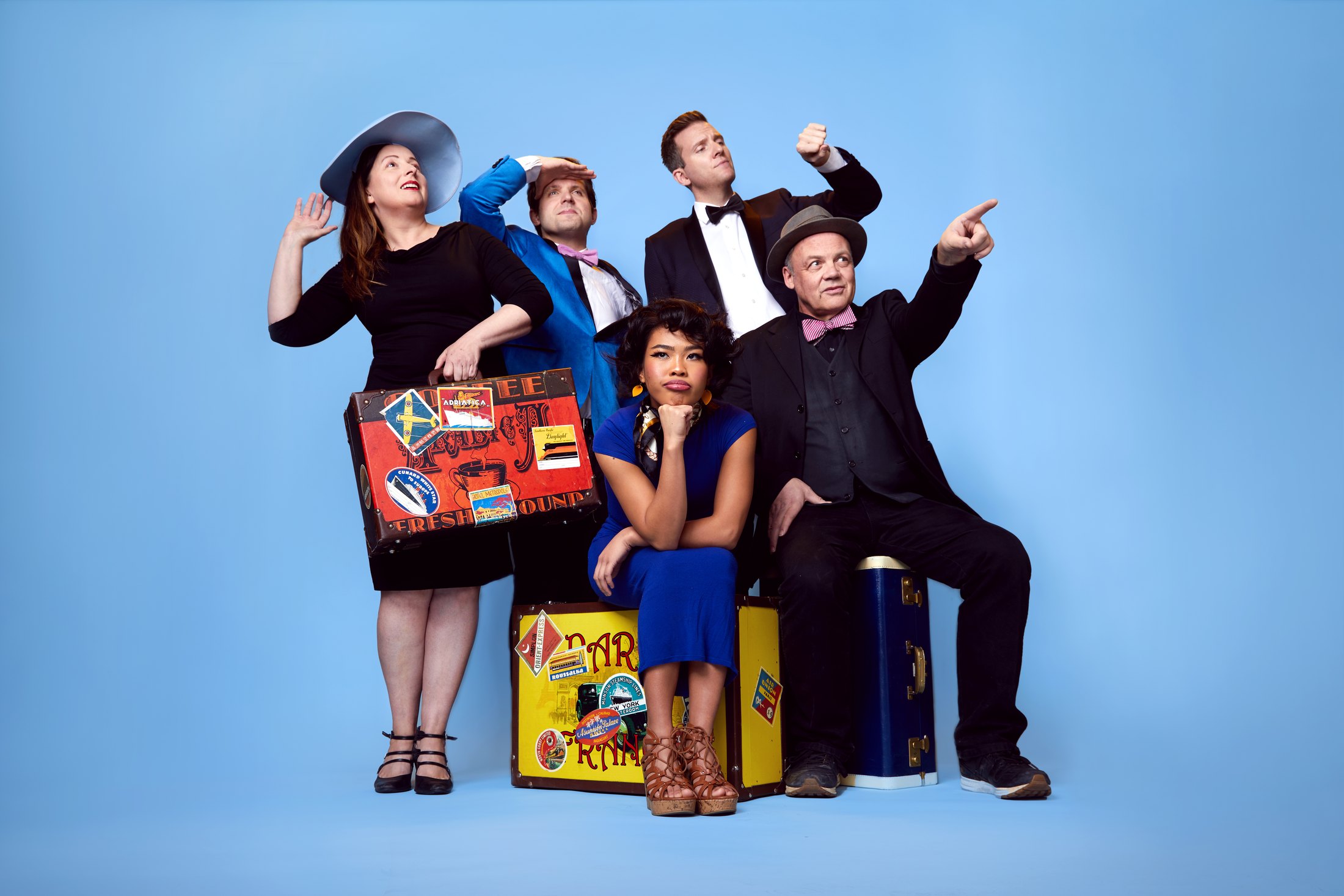 Five performers with vintage suitcases, posed against pale blue