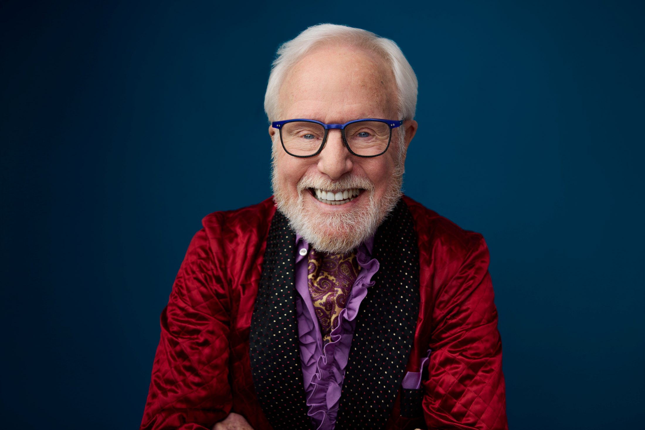 The same man in a red velvet jacket, laughing, against slate