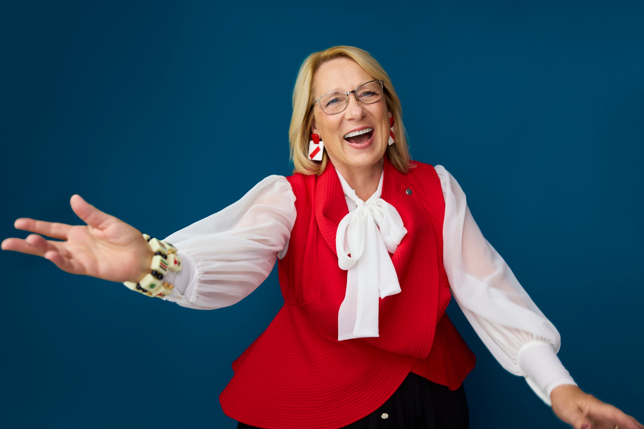 A woman in a red waistcoat and bow blouse, mid laugh, against blue