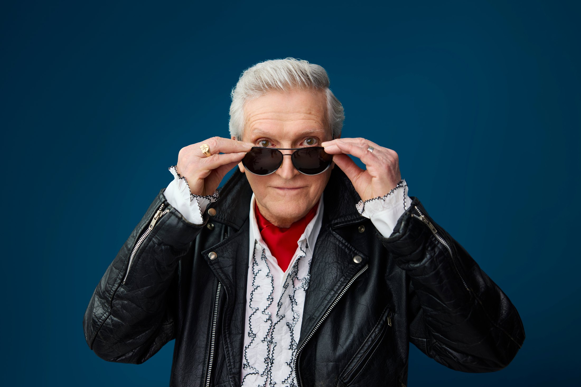 A man in a leather jacket lowering his sunglasses, against teal