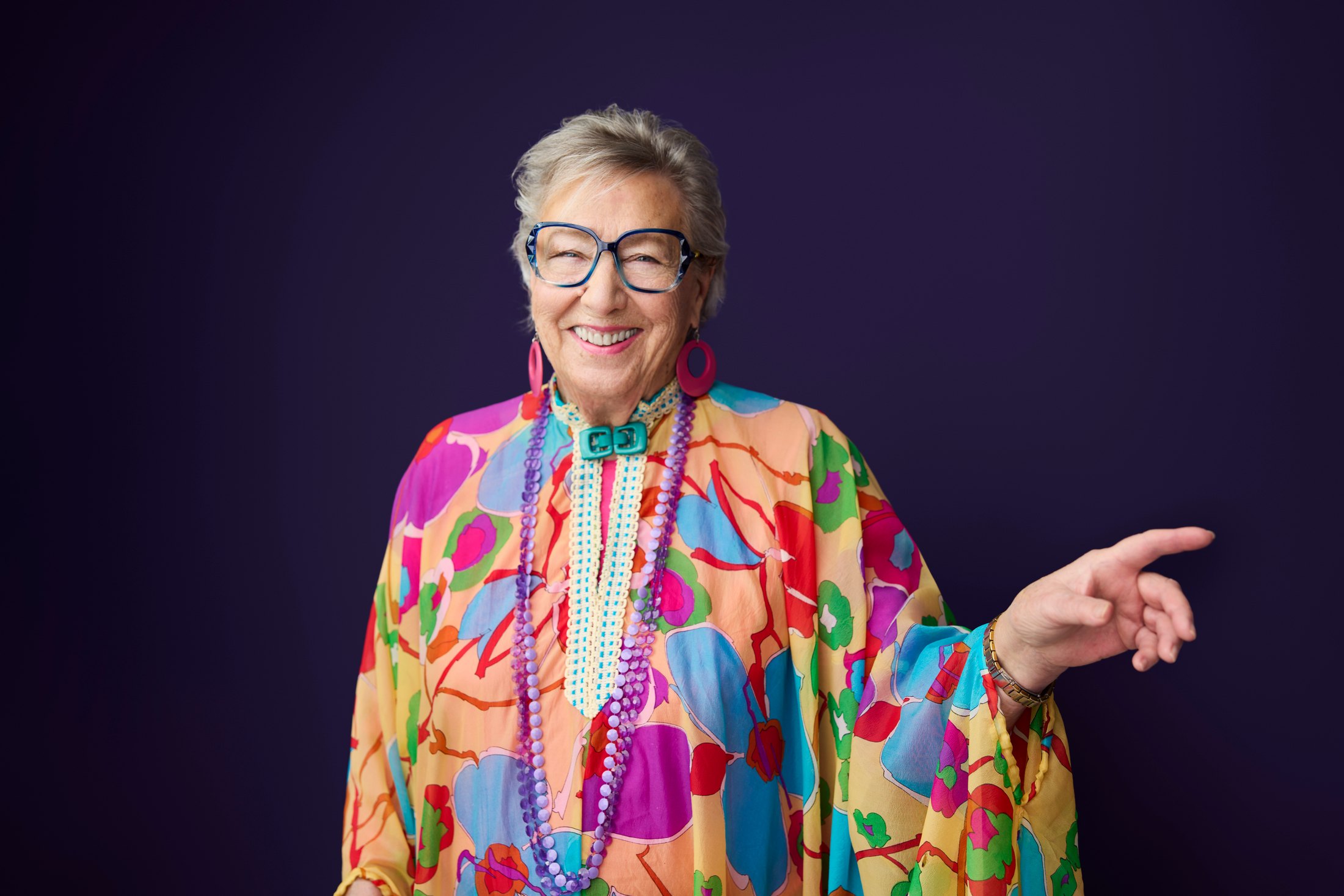 A woman in a bright print top and beads, pointing, against violet
