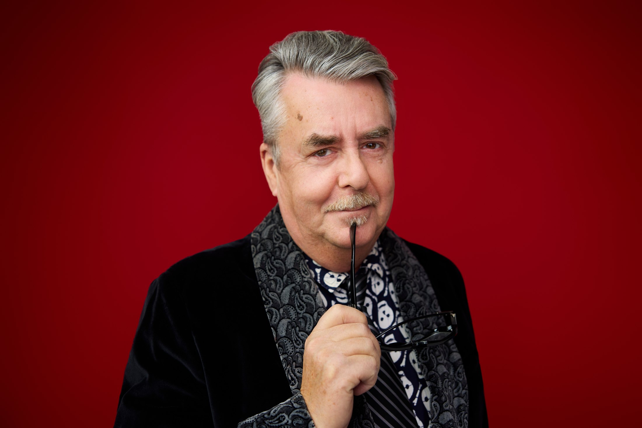 A man in a dark patterned jacket, hand to his chin, against red