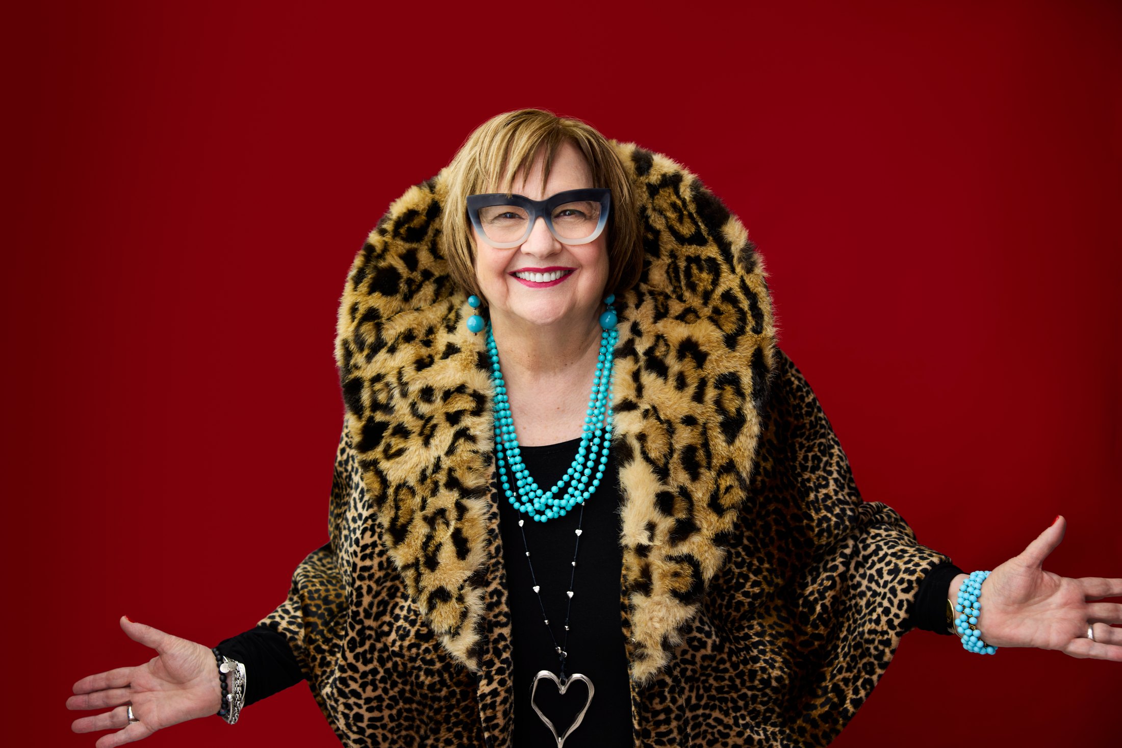 A woman in a leopard-print coat, arms open, against red