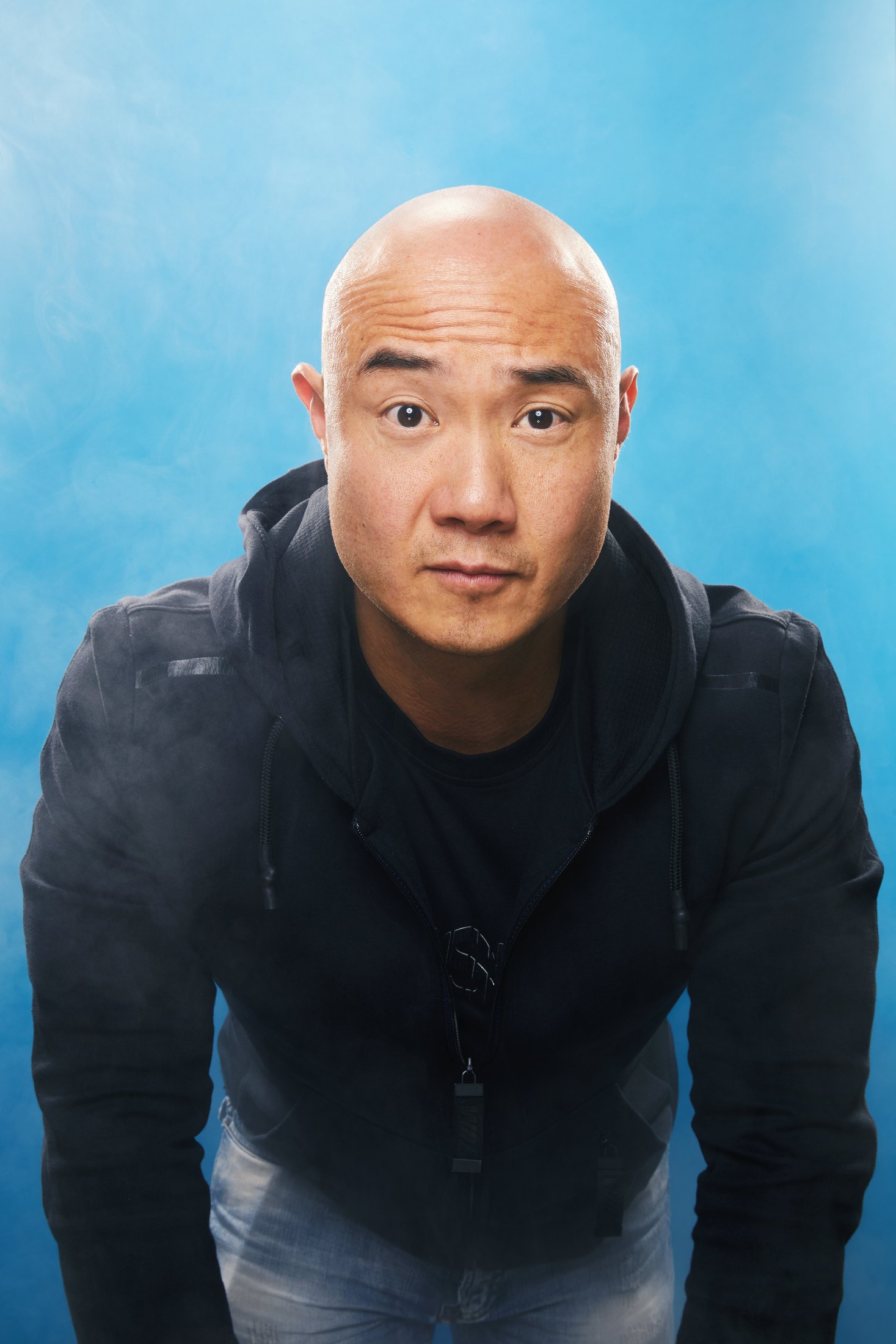 A performer in a dark jacket leaning in toward the camera against pale blue