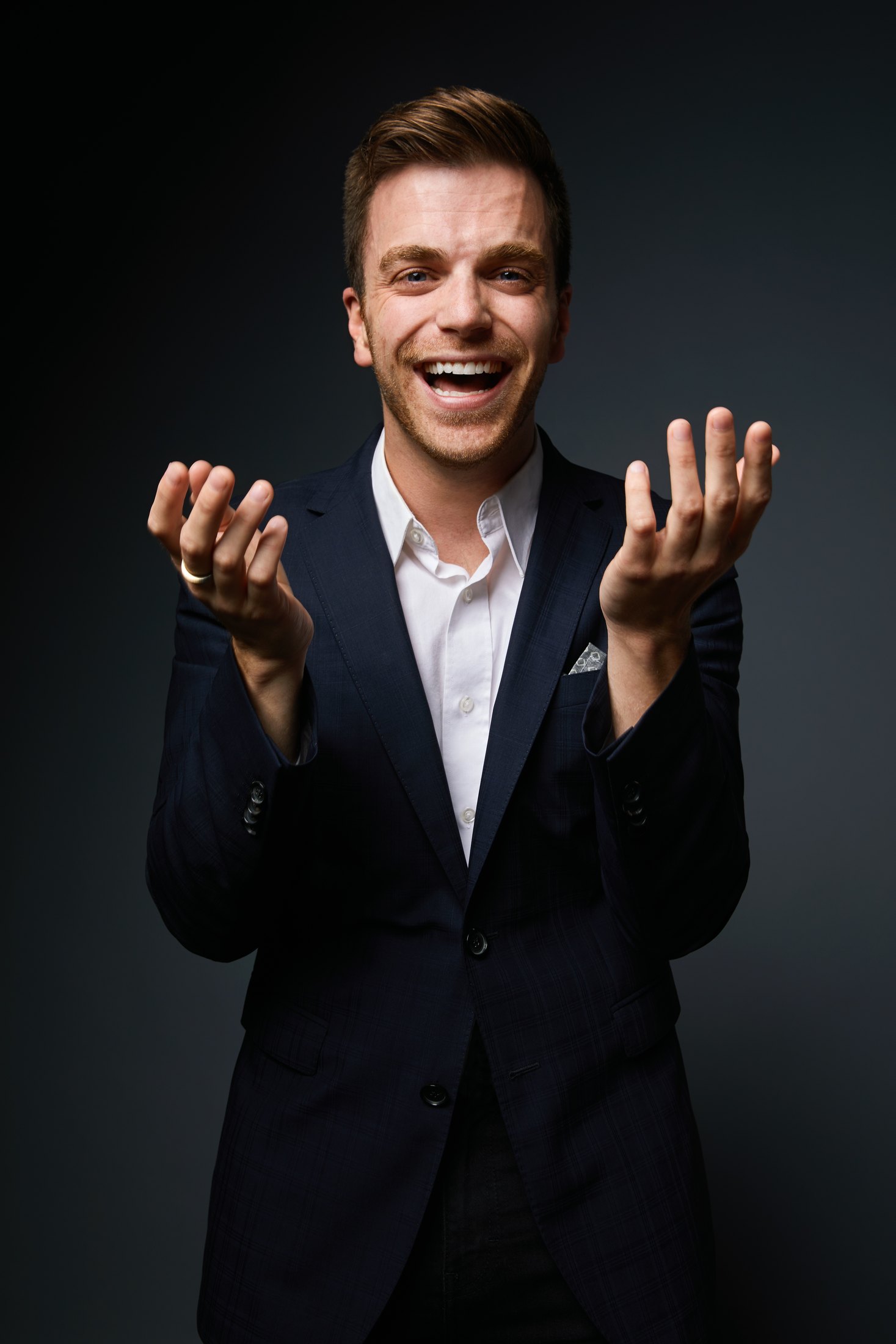 A performer in a dark suit, hands raised, laughing against black