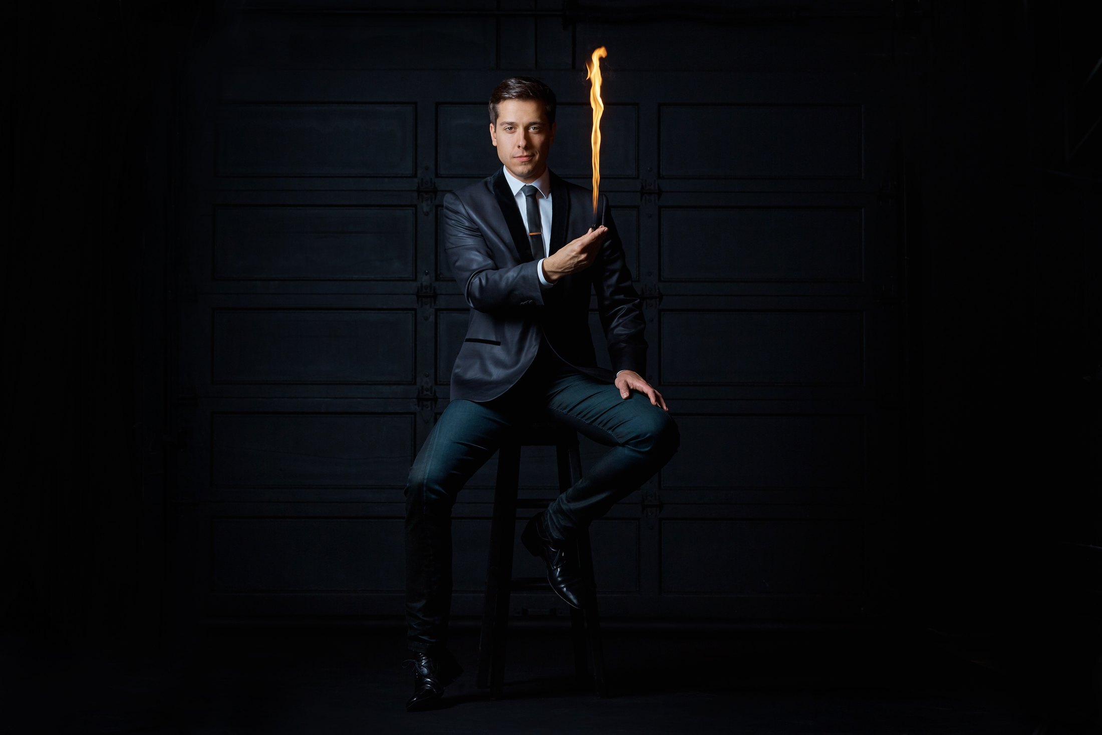 Sheldon Casavant on a stool against black panelled doors, a tall flame rising from his hand