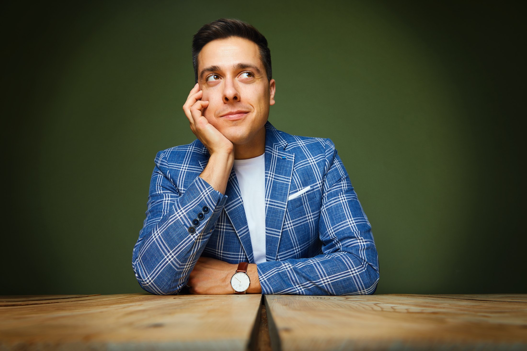 Sheldon Casavant in a blue plaid jacket at a wooden table, chin on his hand, against green