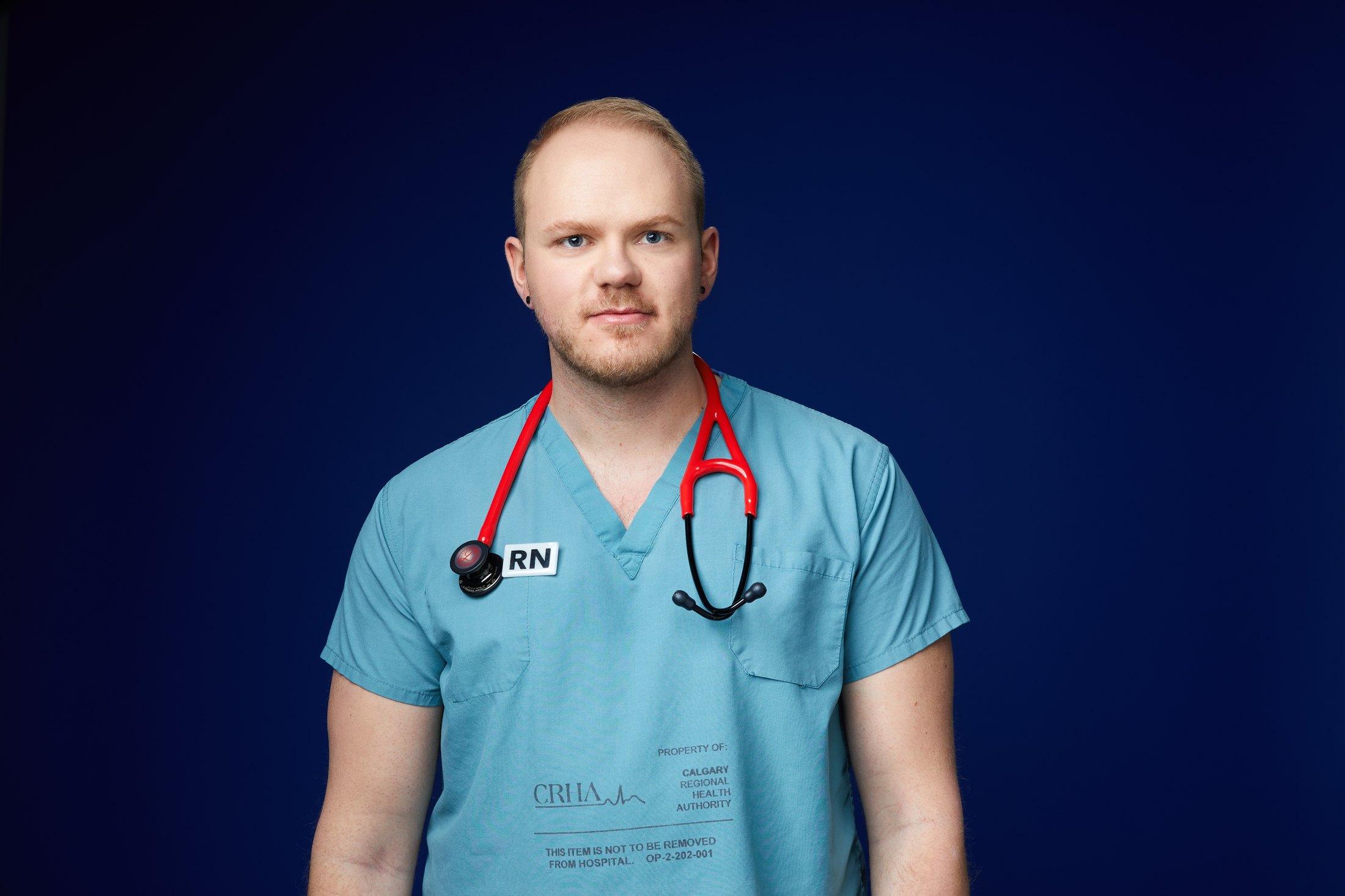A nurse in teal scrubs with a stethoscope around his neck, against deep blue