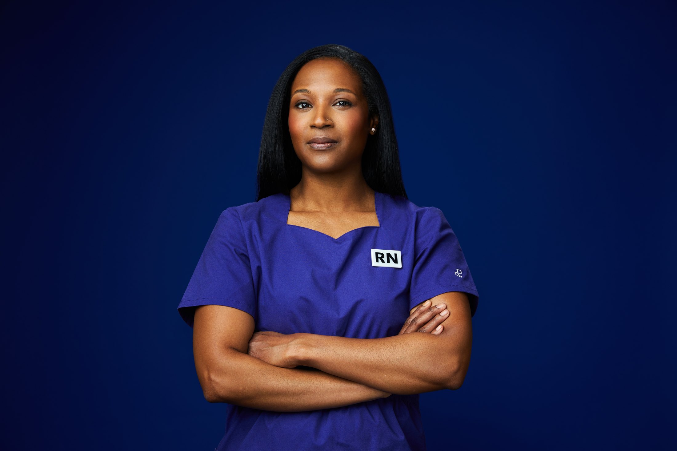 A nurse in indigo scrubs, arms crossed, against deep blue
