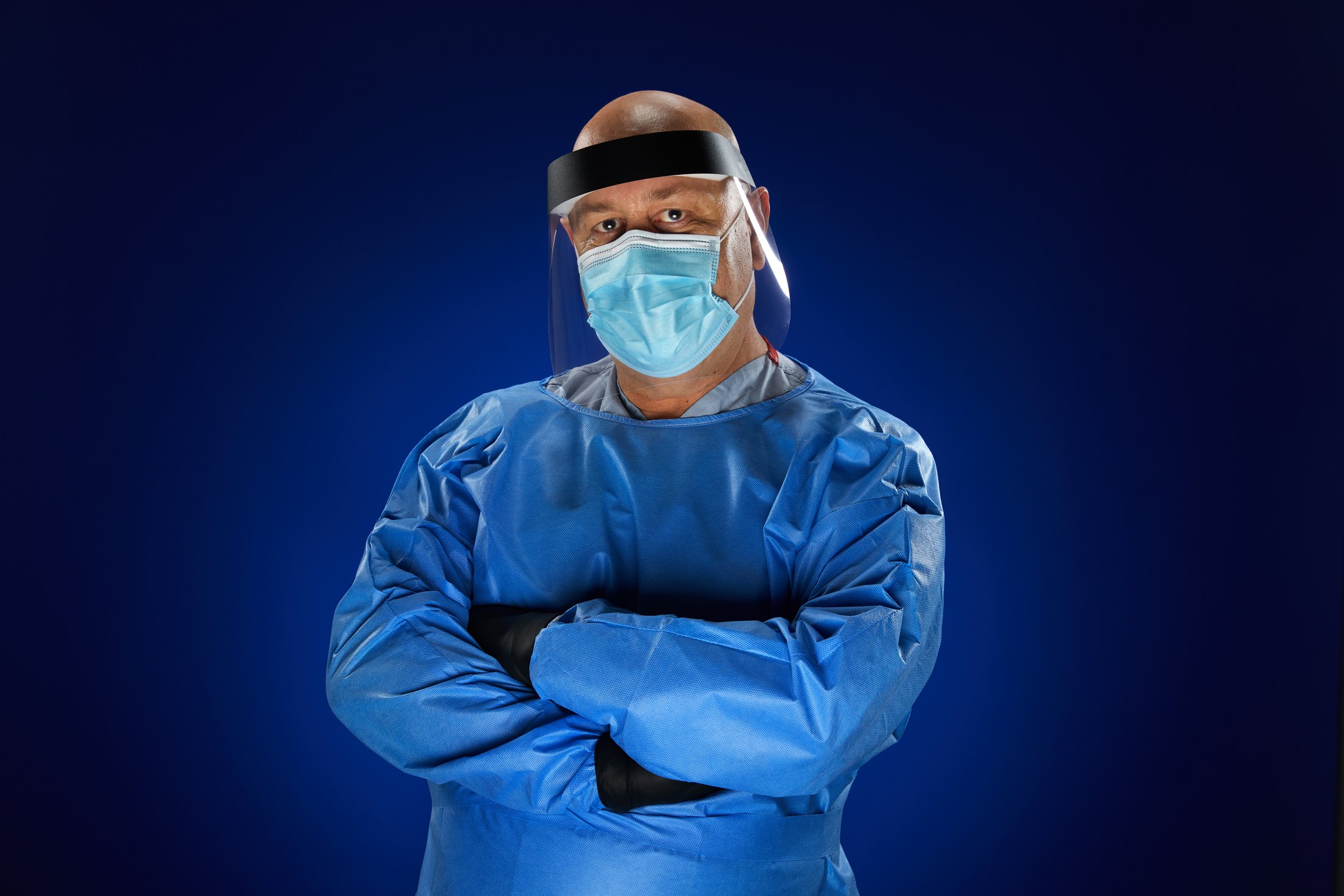 A nurse in a blue isolation gown, face shield and mask, arms crossed, against deep blue