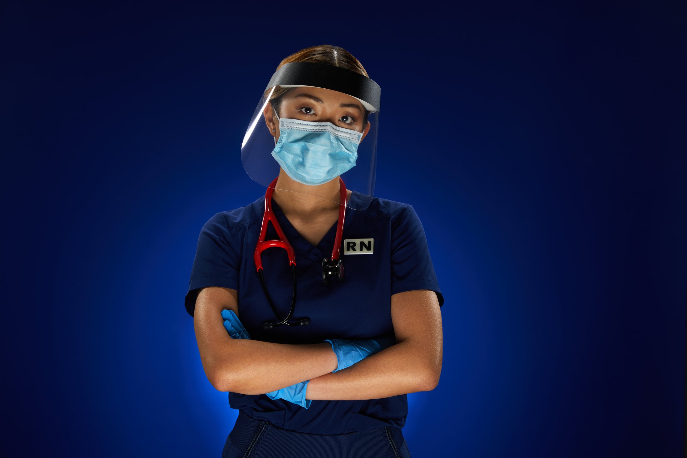 Campaign photography: a nurse in navy scrubs, a face shield and a red stethoscope, arms crossed against deep blue