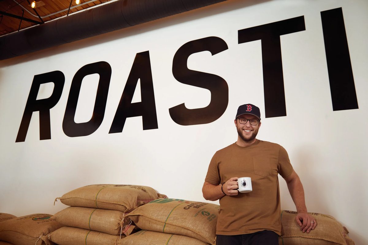 A man with a coffee cup leaning on stacked burlap sacks beneath a painted ROASTI wall sign