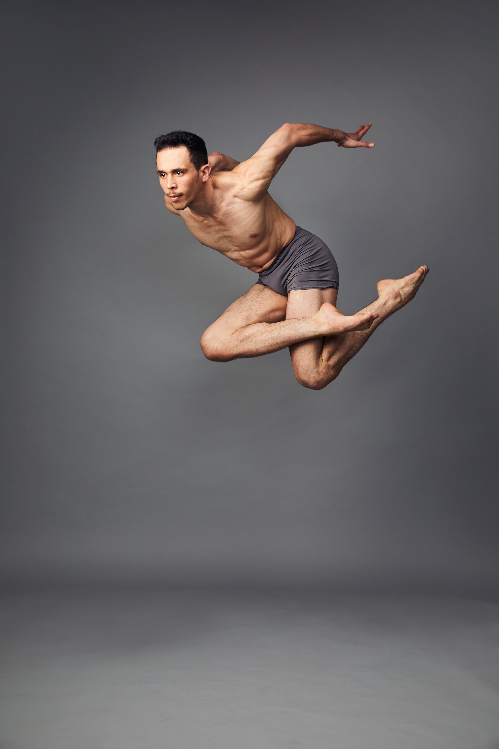 A dancer in grey shorts mid-leap on grey, knees bent under him, one arm swept back
