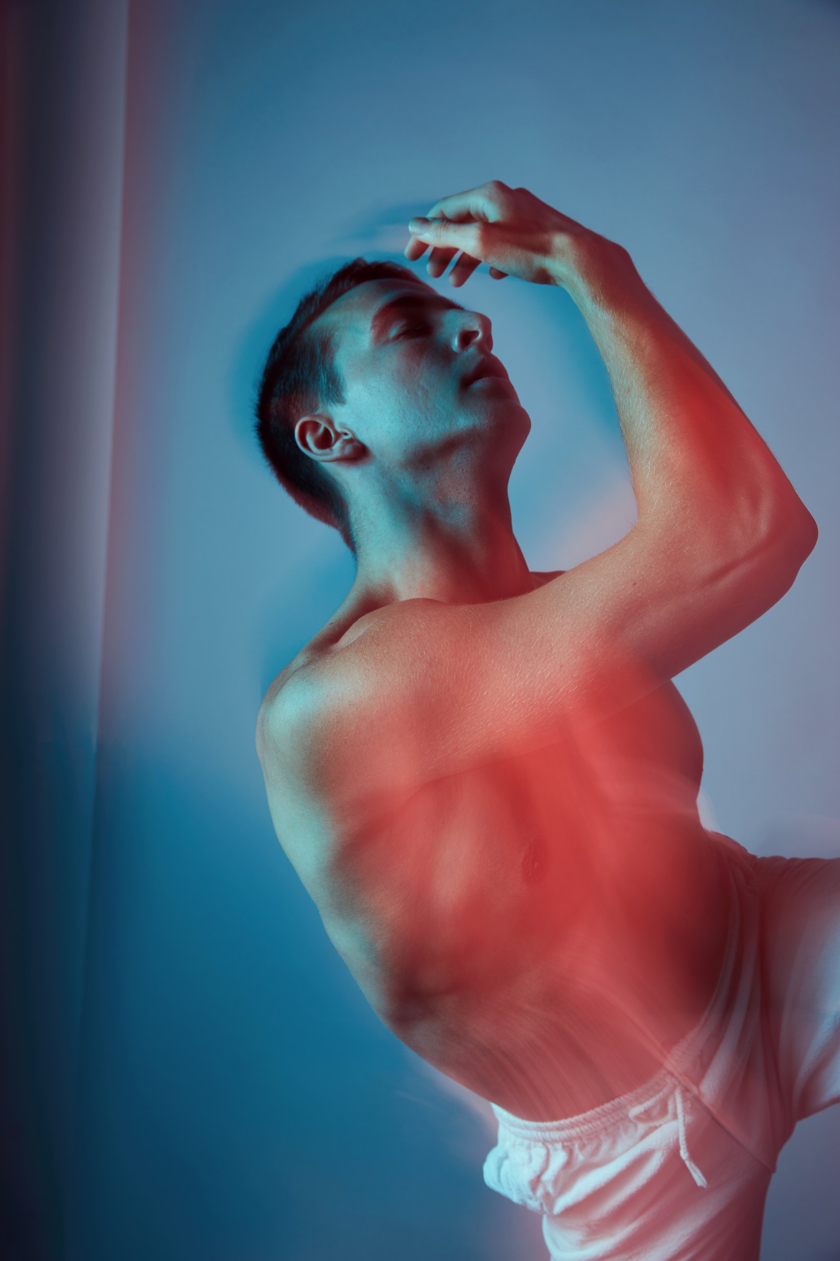 Under red and blue gels, torso turned, one hand raised to his brow, looking up