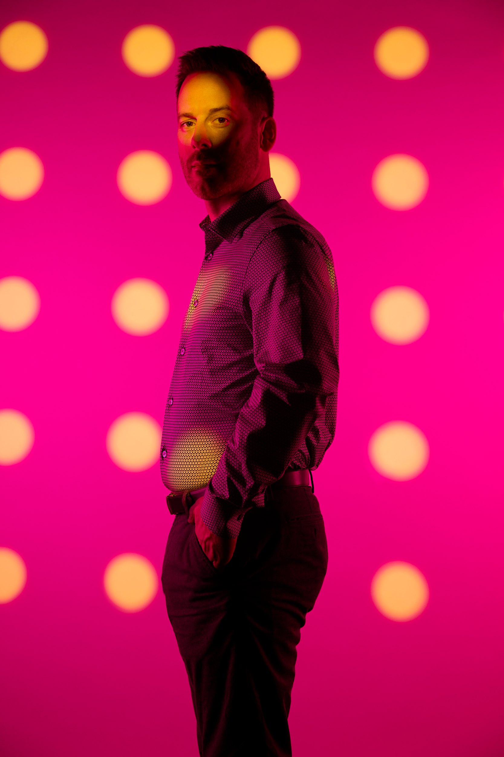 Standing, hands at his sides, the dots across the wall behind and across him