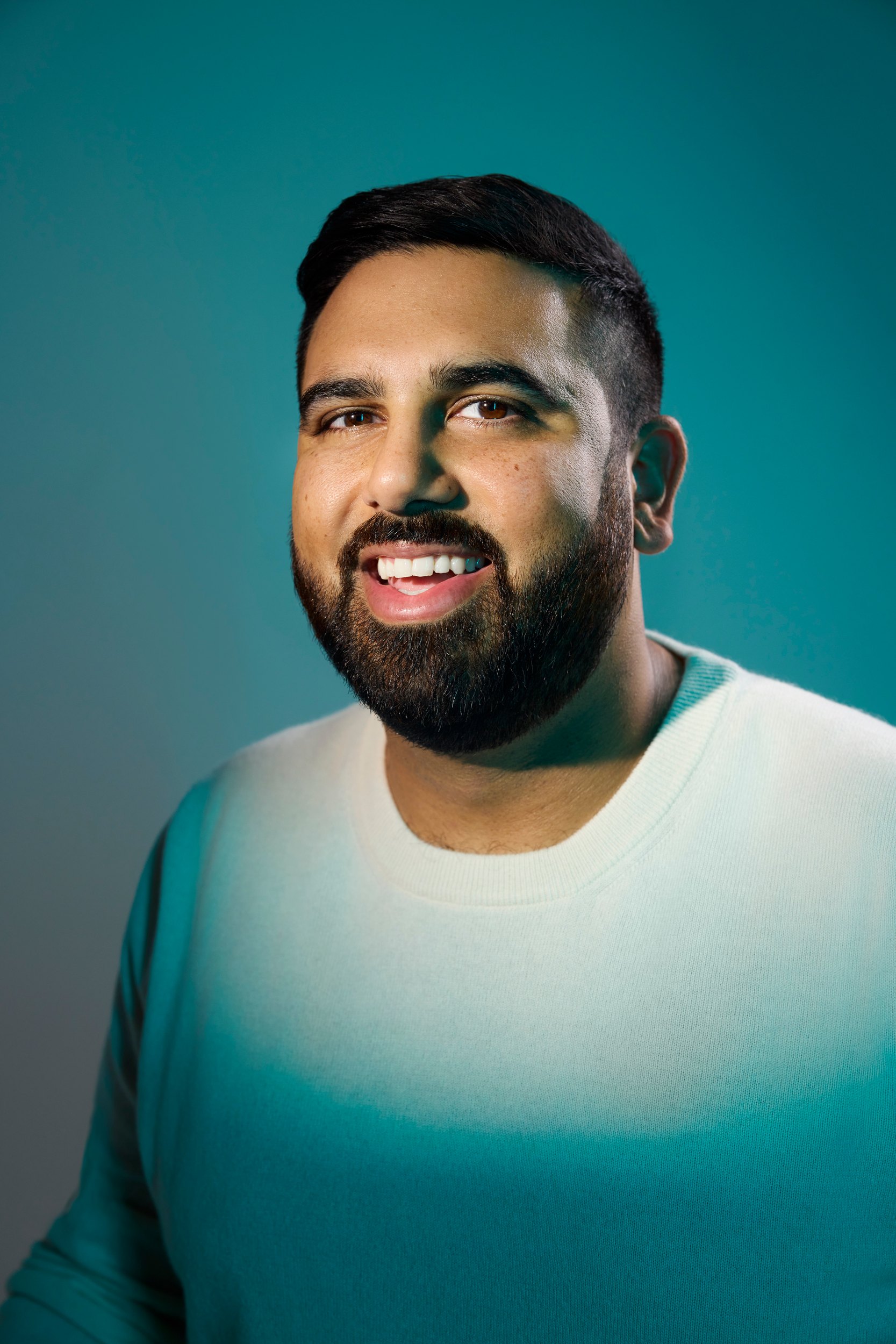 Sukhwinder Johal in a pale green sweatshirt against teal, mid laugh