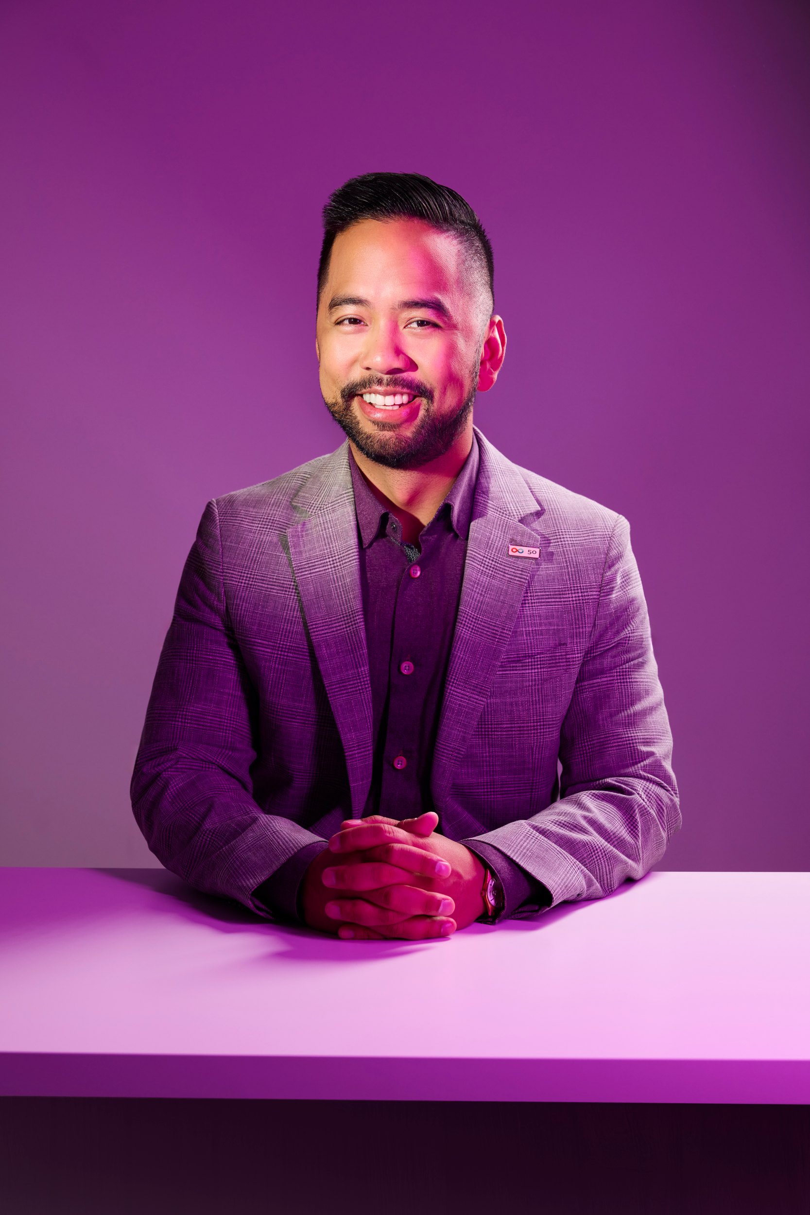 Angelo Talamayan in a pale suit at a lilac table, hands folded