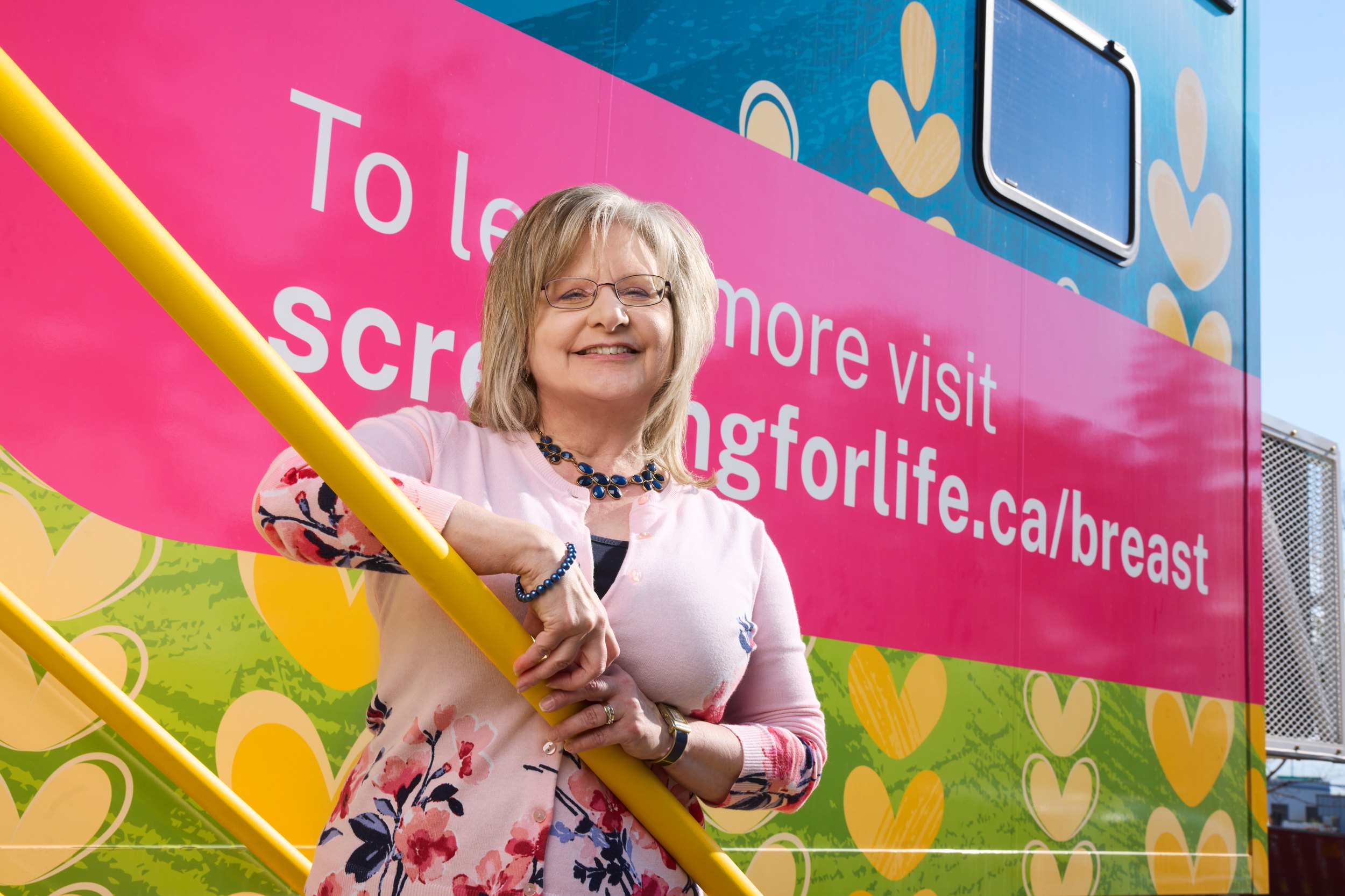 Joan Hauber leaning on the yellow rail of a pink breast screening bus, smiling