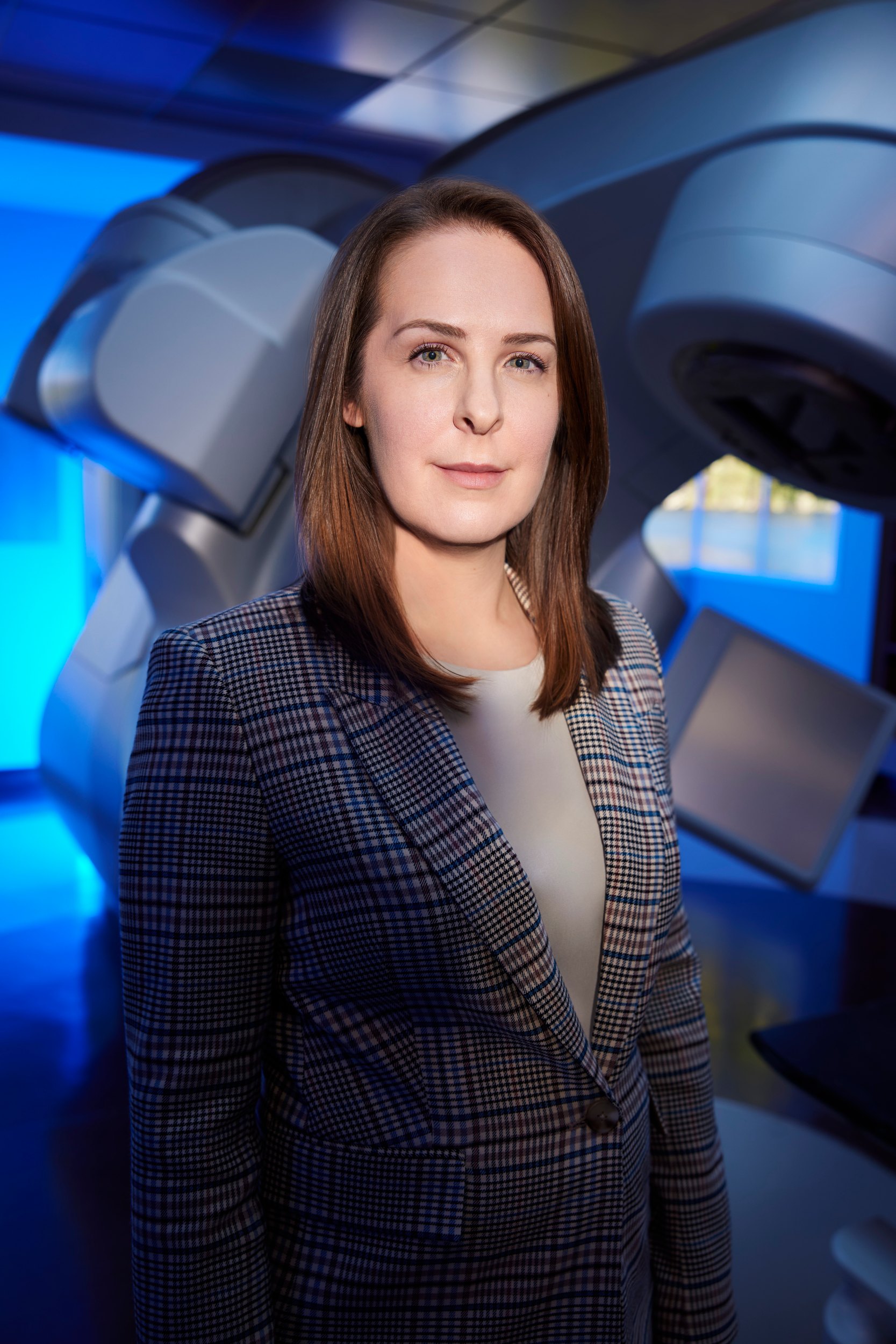 The physician against the machine, upright, the blue of the suite behind her