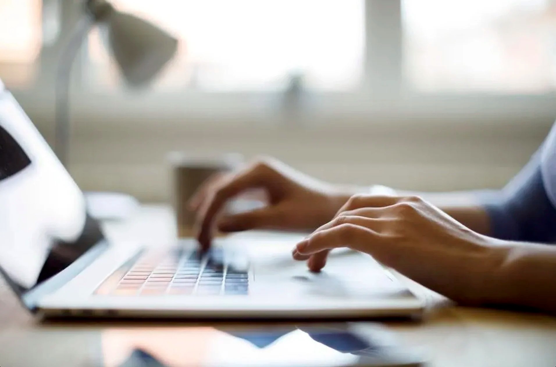 Close-up of person's hands typing on a laptop keyboard in a bright room.