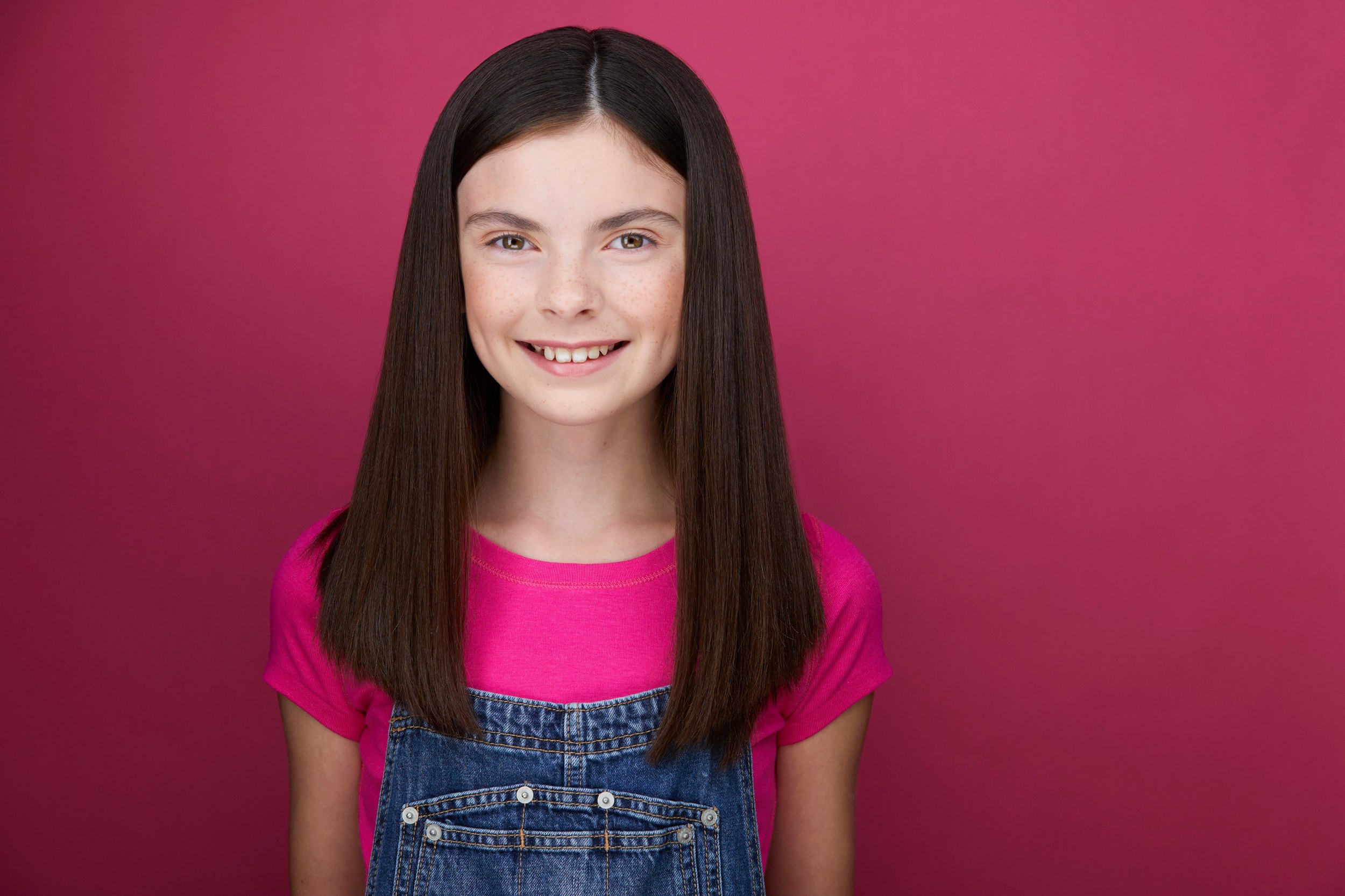 The same session, a pink top and overalls against pink, smiling