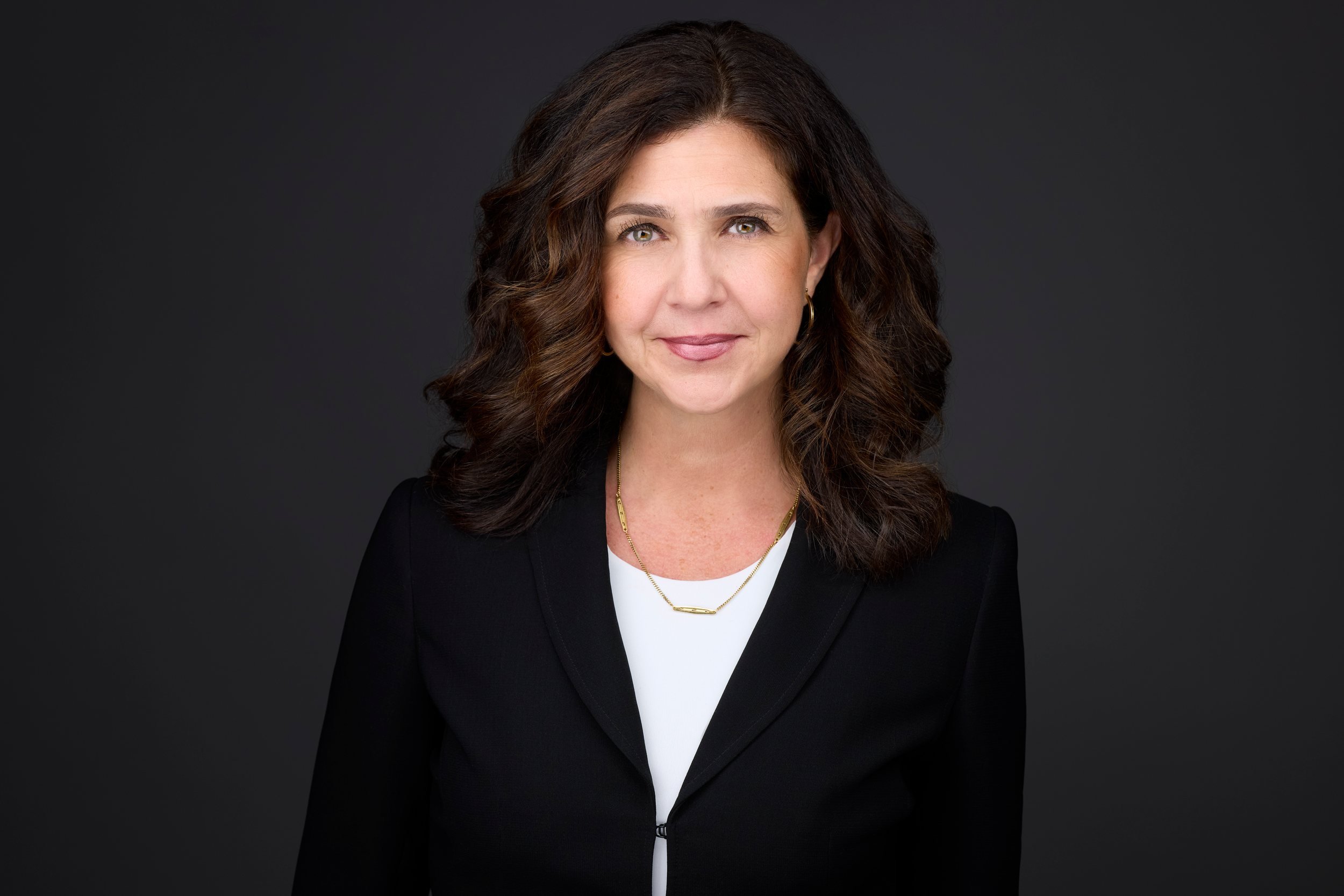 Real estate headshot: dark curls, a black blazer and gold necklace, on dark grey