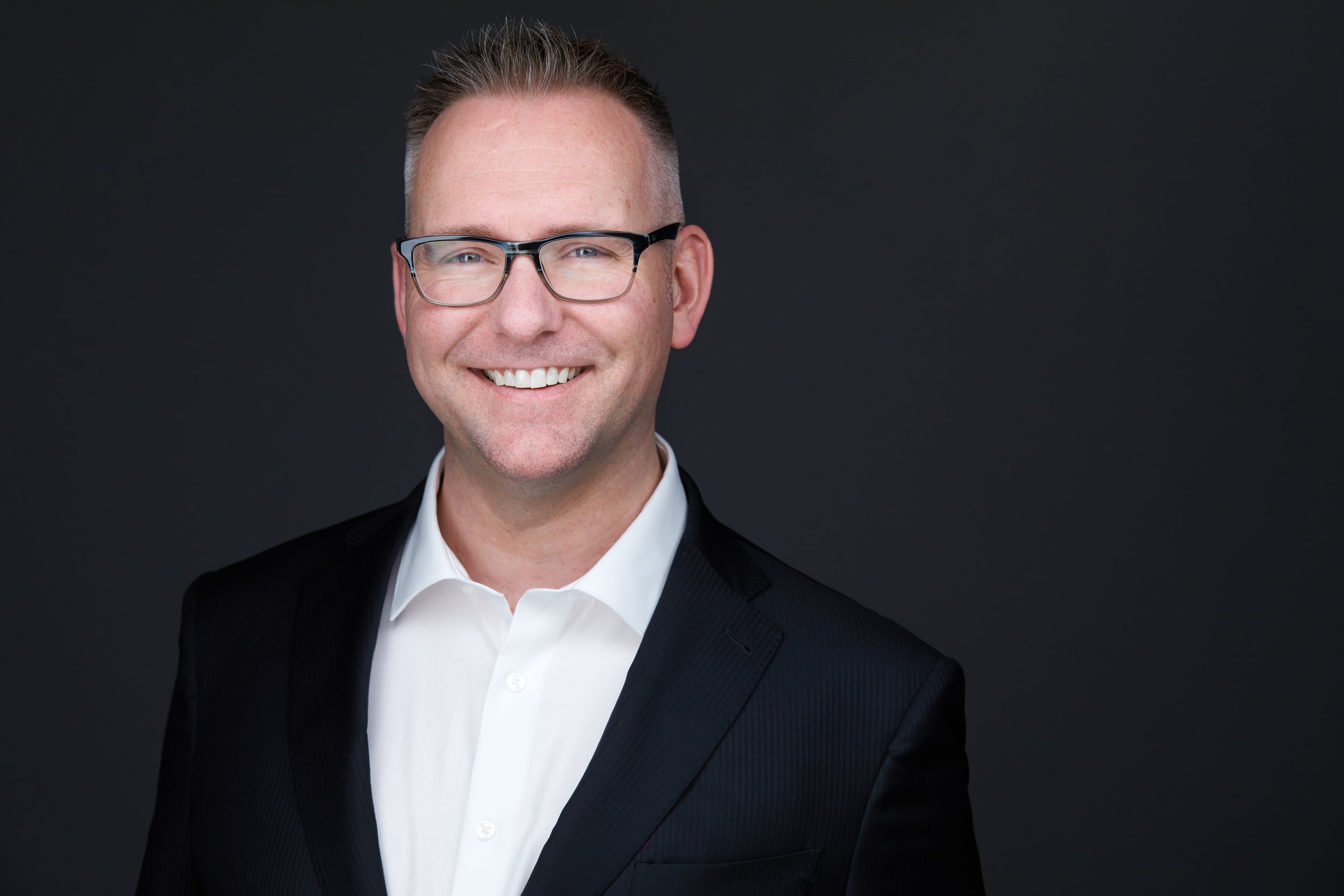 Real estate headshot: glasses, a black blazer and open collar, on dark grey