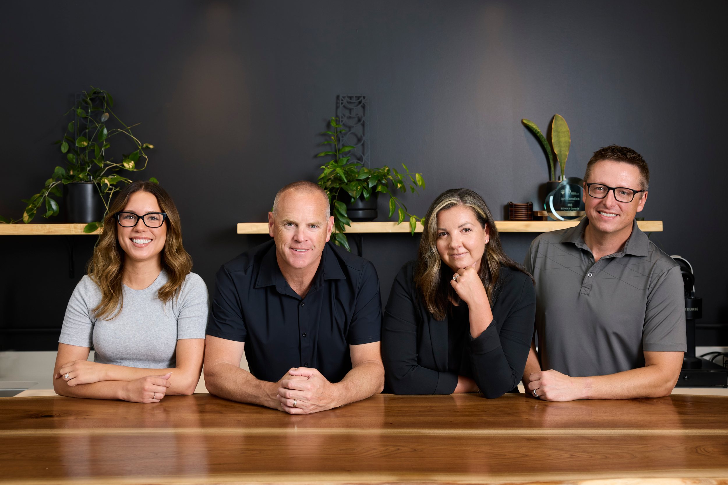 A real estate team of four at a long wooden table in their own office, a black wall and shelves of plants behind them