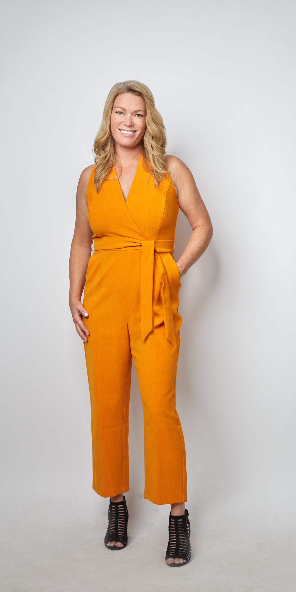 Full-length portrait on light grey: an orange jumpsuit tied at the waist and heeled sandals