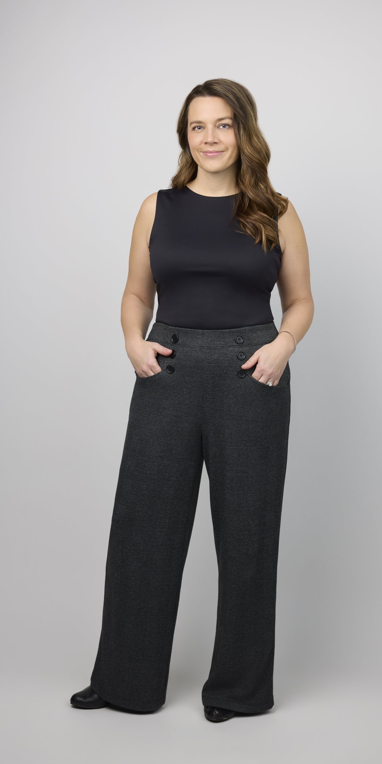 Full-length portrait on light grey: a black sleeveless top and wide charcoal trousers, hands in pockets