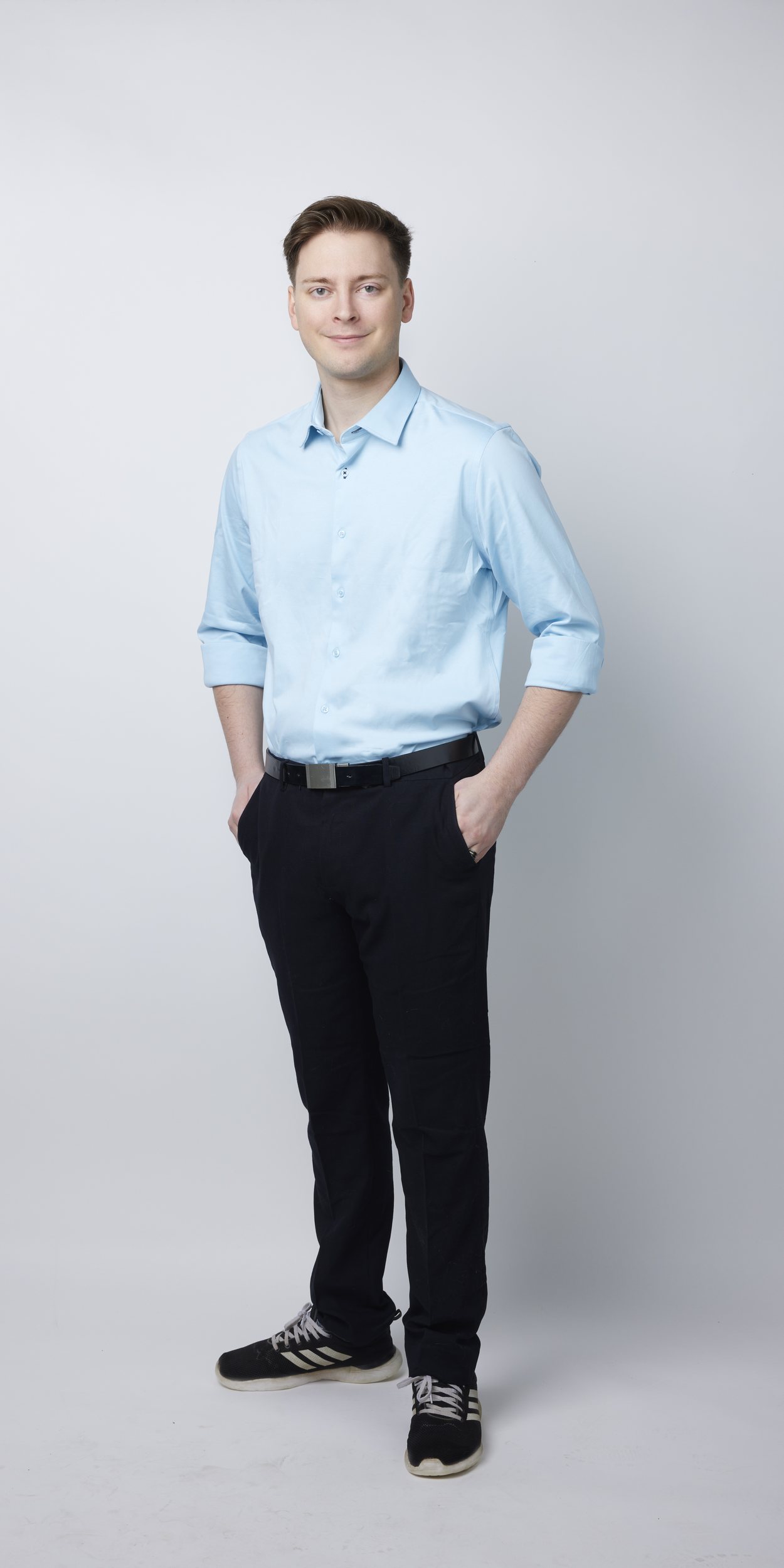 Full-length portrait on light grey: a light blue shirt with the sleeves rolled, hands in pockets