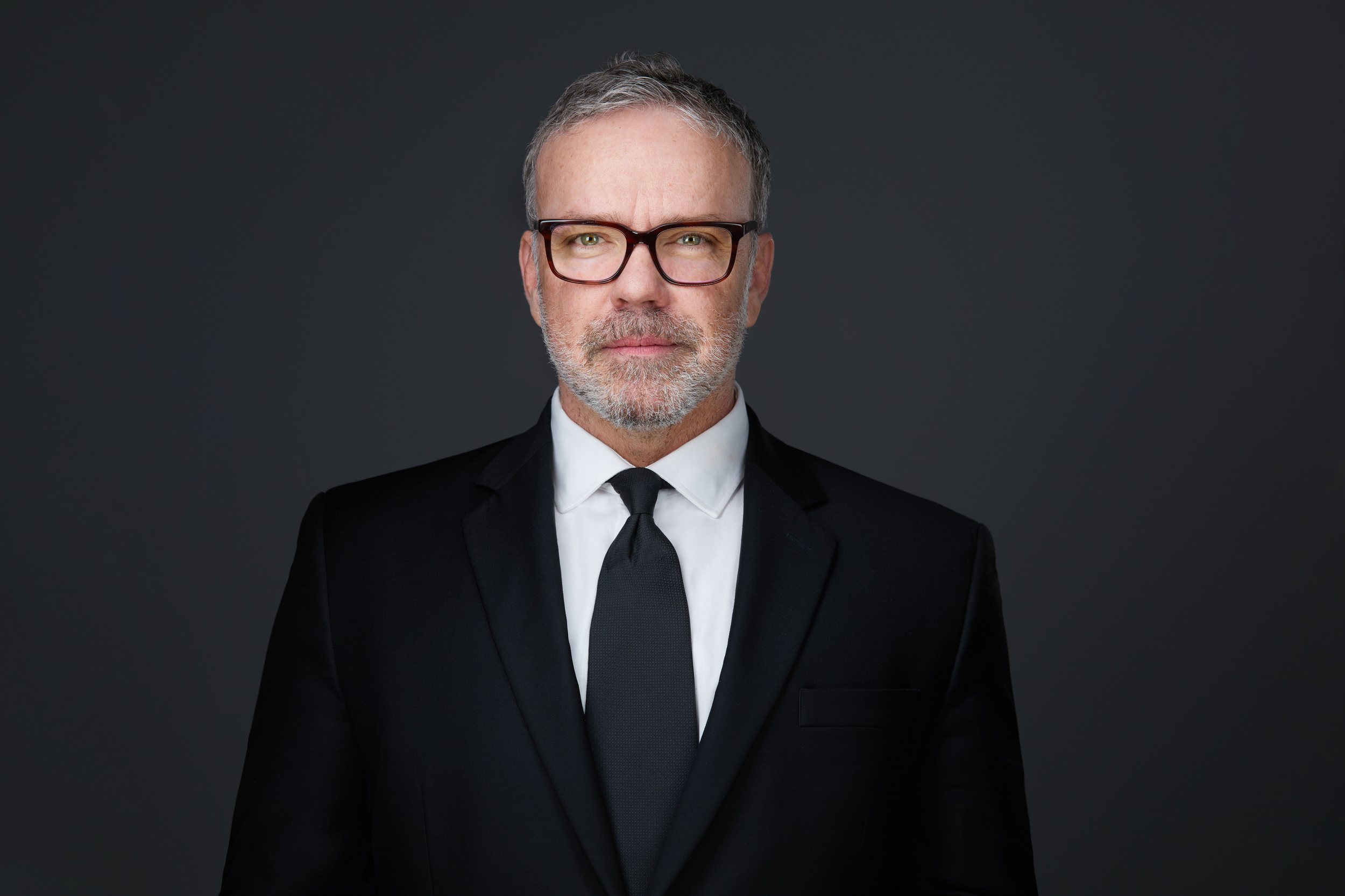 Real estate headshot: glasses and a grey beard, a black suit and tie, on dark