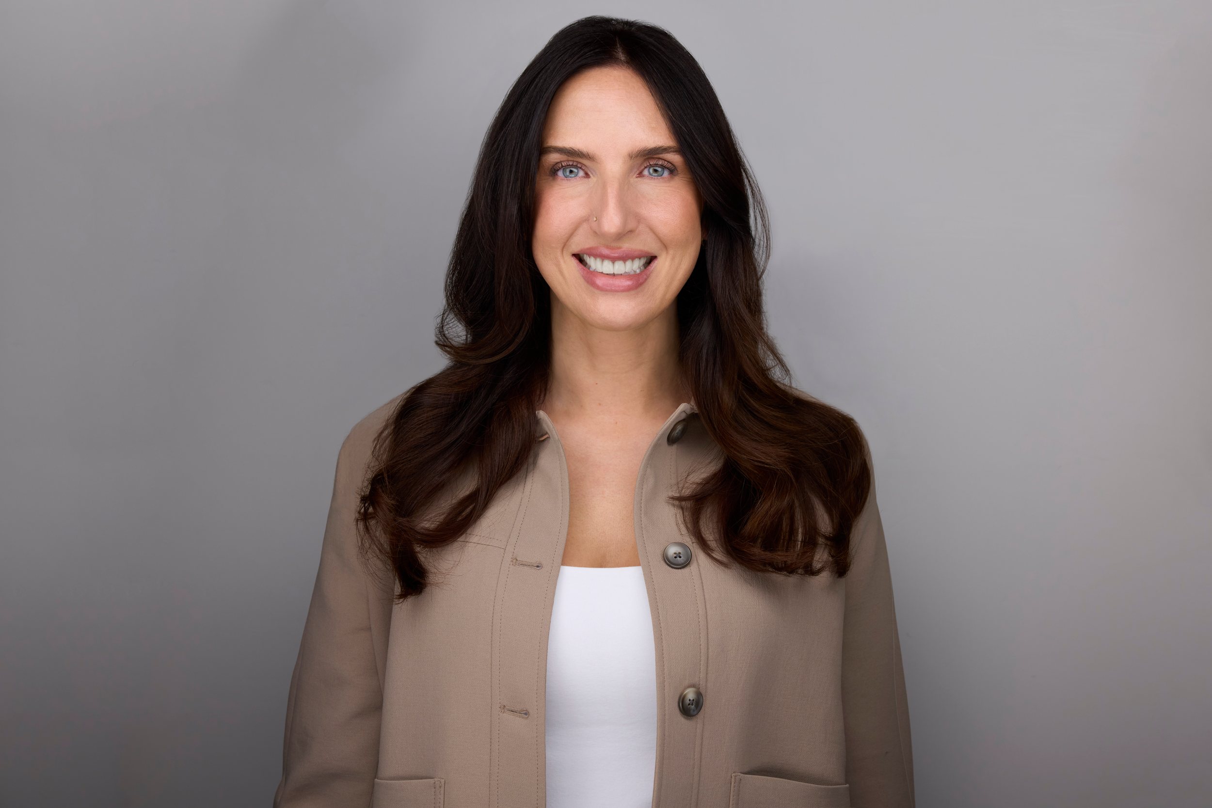 Real estate headshot: long dark hair, a tan jacket over white, on light grey