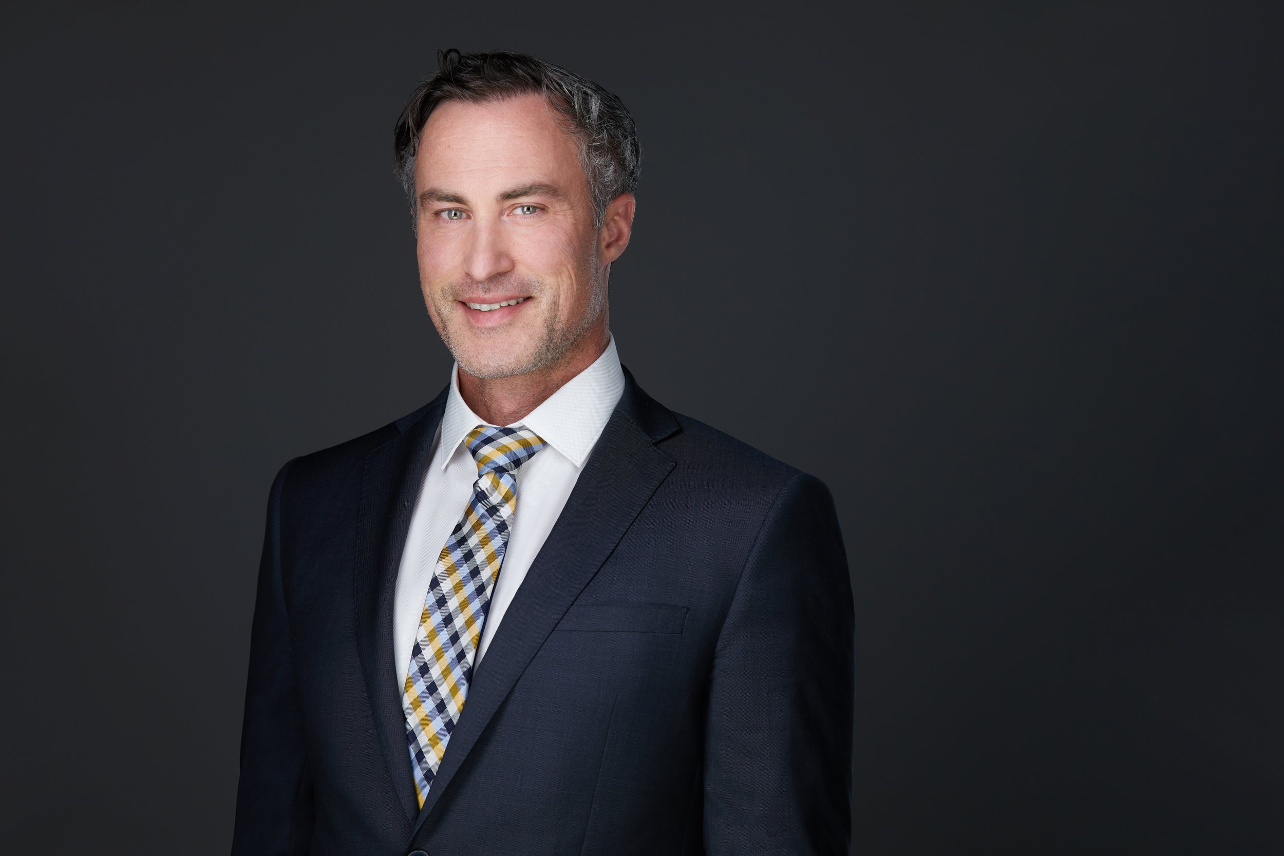 Real estate headshot: a navy suit and a patterned tie, smiling, on dark grey