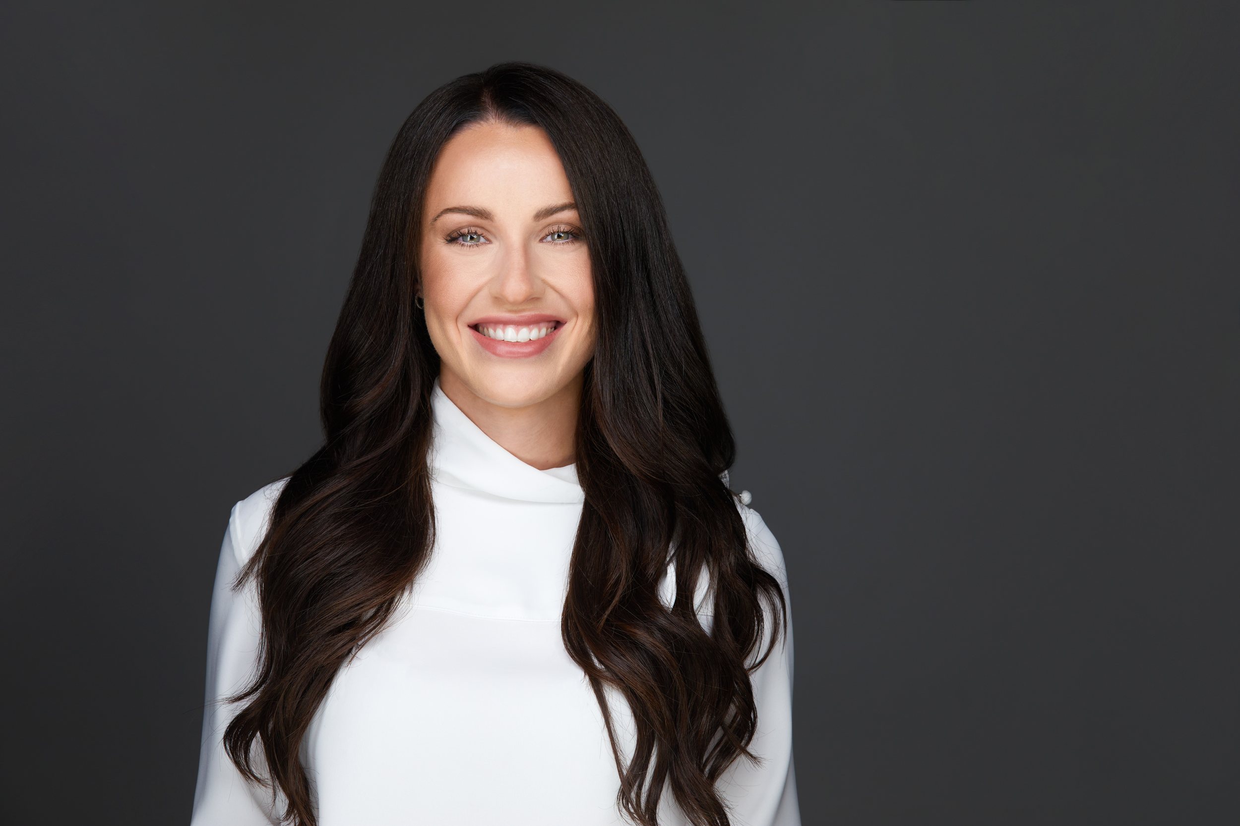 Real estate headshot: long dark waves, a white high-neck top, on dark grey