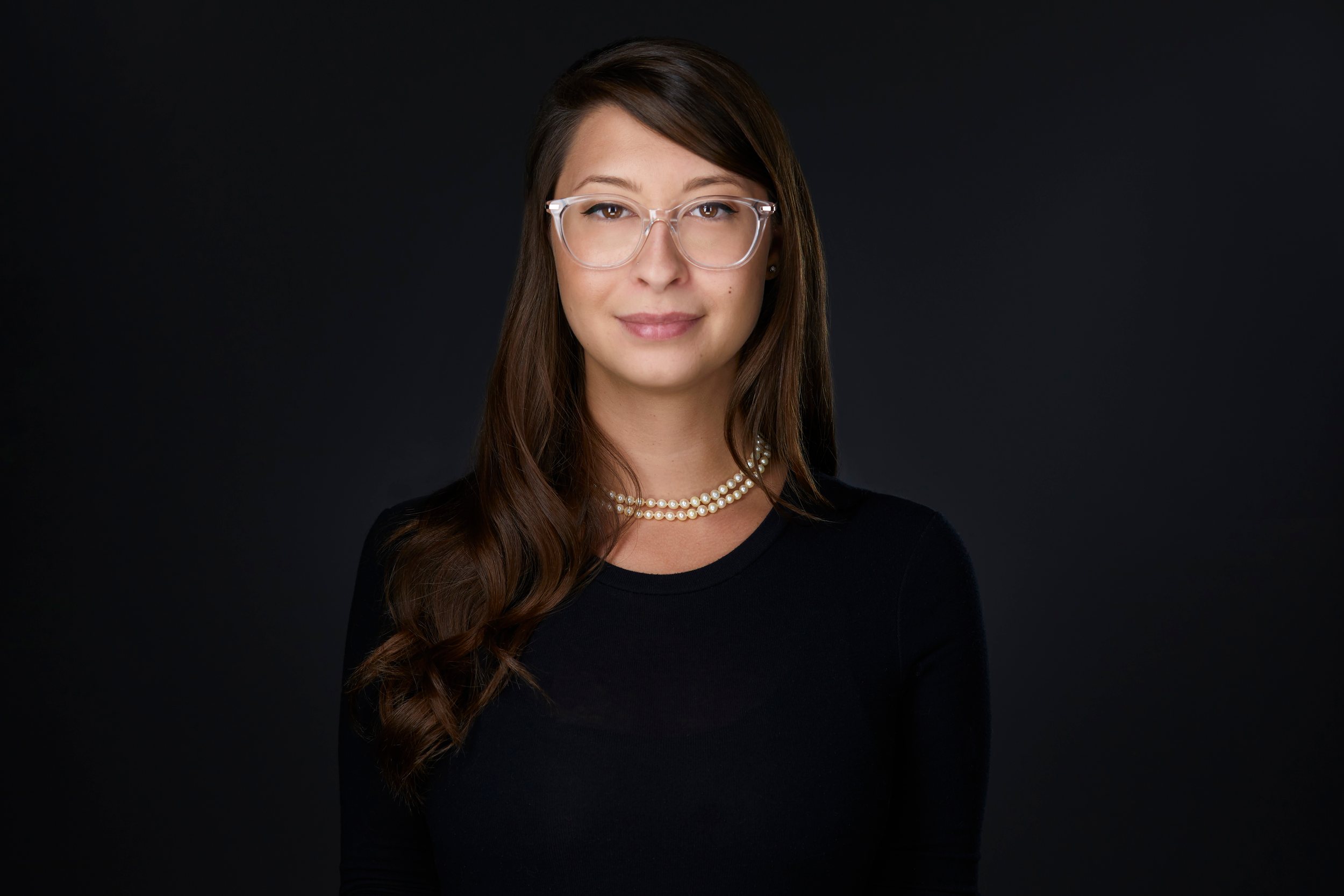 Real estate headshot: glasses, long dark hair, a black top and pearls, on dark