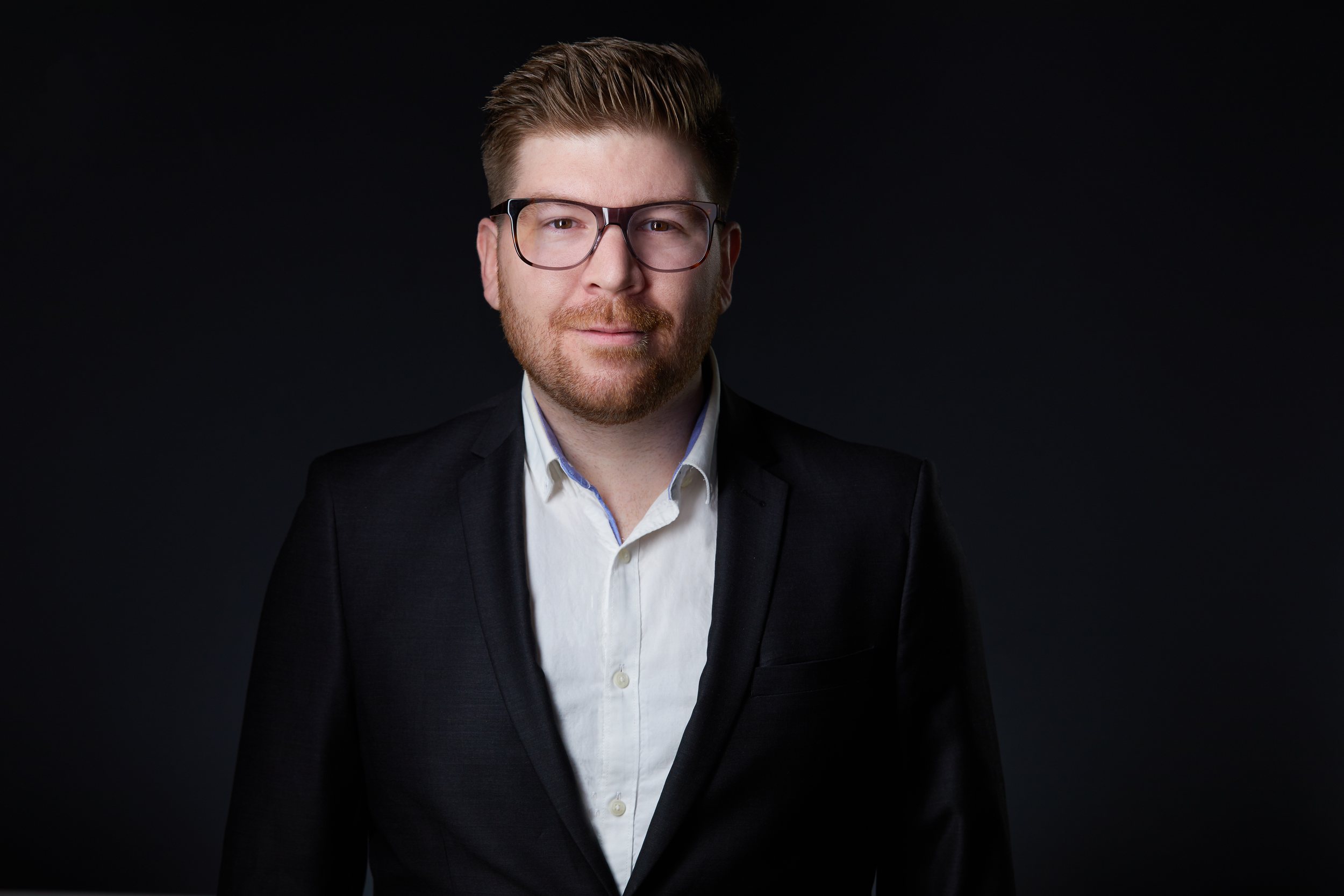 Real estate headshot: glasses, a beard, a black suit and open white collar, on dark