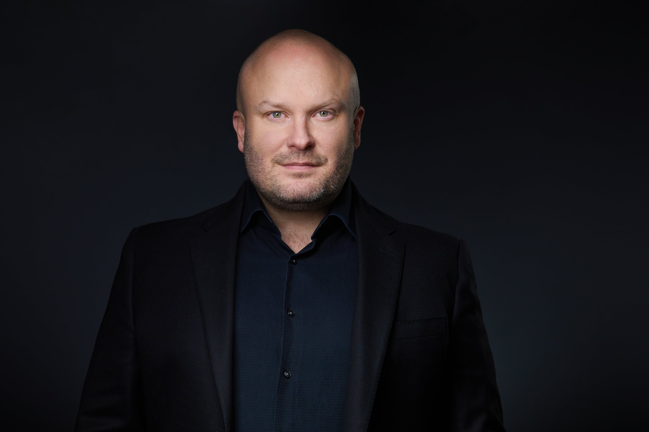 Real estate headshot: a shaved head, a black suit over a black shirt, on dark