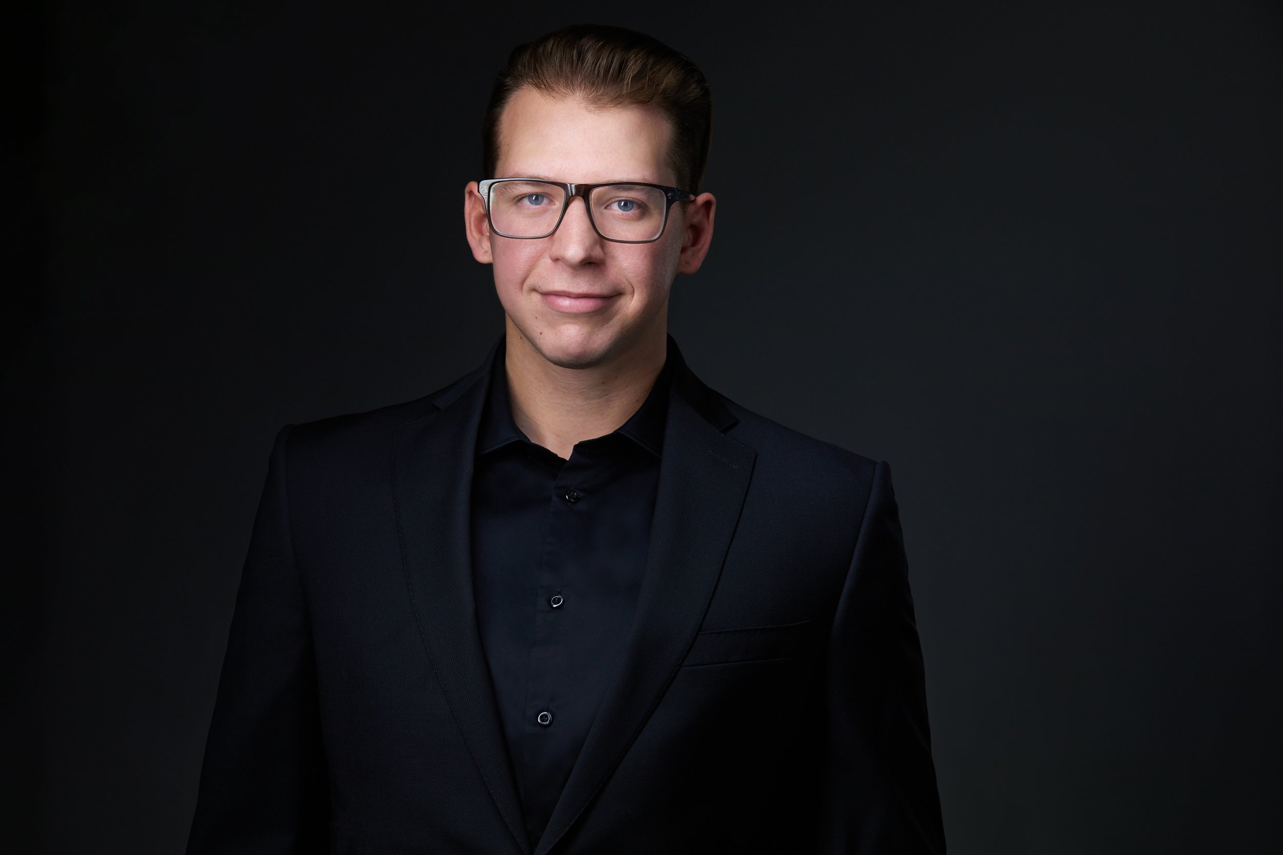 Real estate headshot: glasses, a black suit over a black shirt, on dark