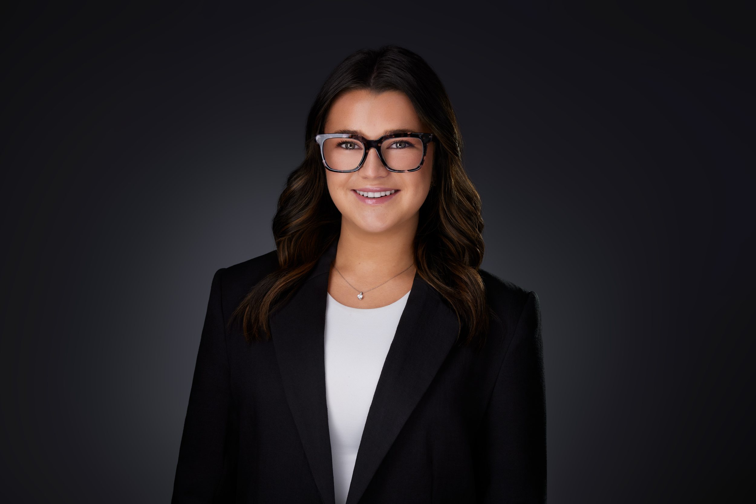 Real estate headshot: glasses, long dark hair, a black blazer over white, on dark